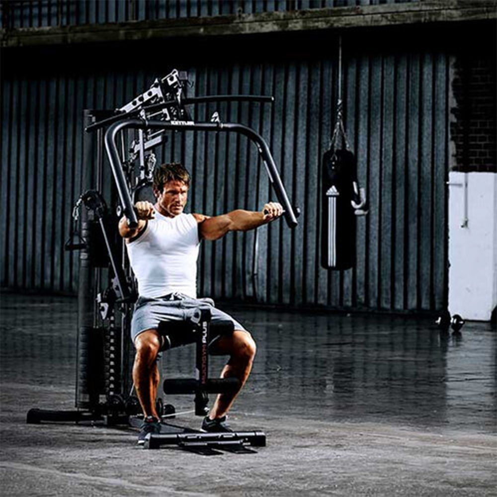 Raumeningas vyras, apsirengęs baltus marškinėlius be rankovių ir pilkus šortus, atlieka krūtinės pratimus ant jėgos staklių KETTLER MULTIGYM PLUS, esančių sporto salės aplinkoje