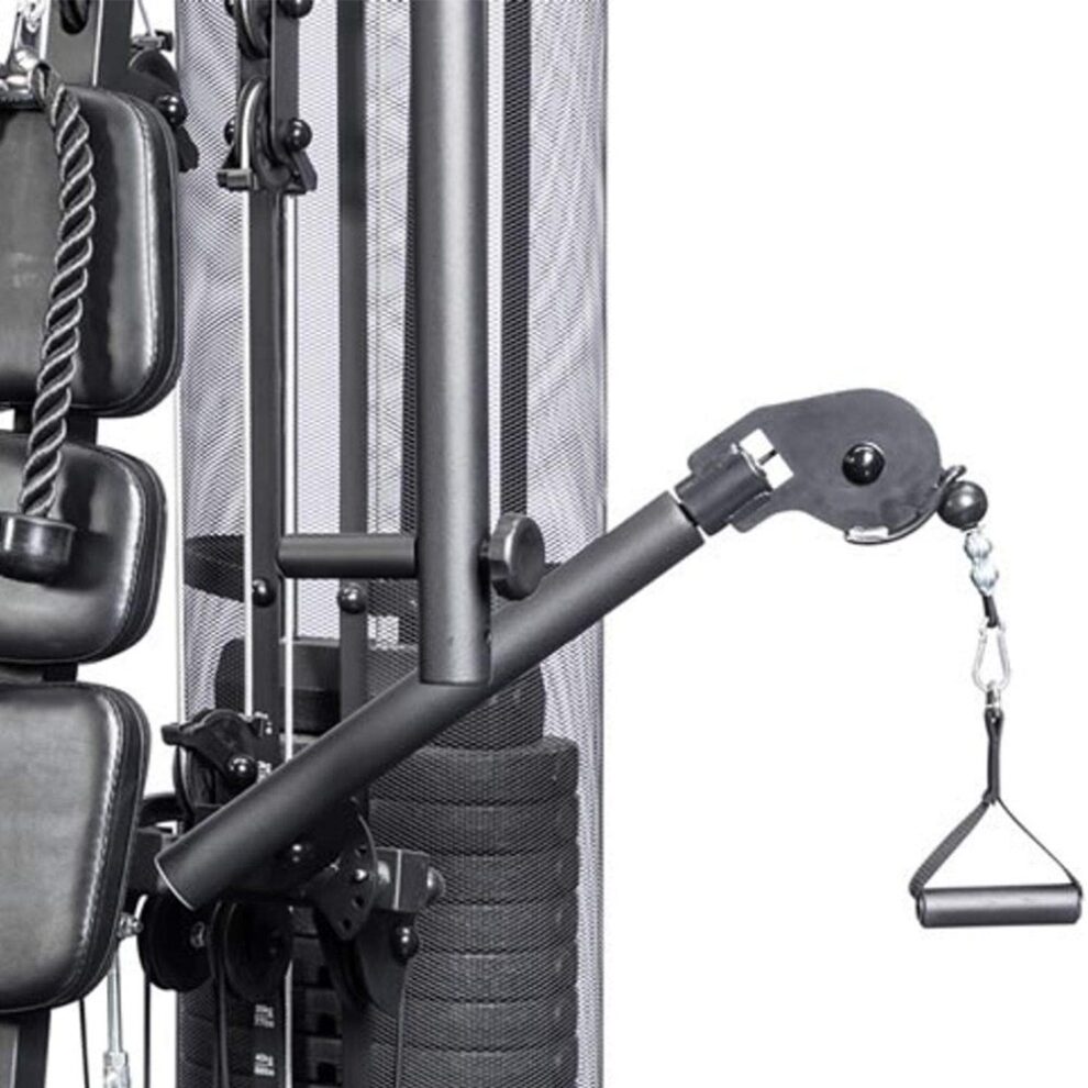 Stambiu planu nufotografuotos juodos spalvos jėgos staklės KETTLER MULTIGYM PLUS 4, rodančios trosų sistemą ir svarmenų bloką su tinkliniu apsauginiu gaubtu